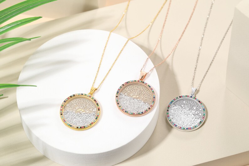May include: Three pendant necklaces with round, tree-of-life designs. One is gold-toned, one rose gold, and one silver. Each pendant has a colorful gemstone border and hangs on a delicate chain. The necklaces are displayed on a white, circular platform.