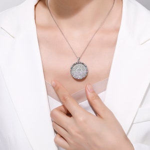 May include: A silver pendant necklace with a round, ornate design. The pendant is encrusted with numerous small, sparkling stones, creating a dazzling effect. The necklace is worn against a white blazer.