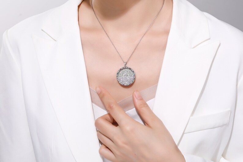 May include: A silver pendant necklace with a round, ornate design. The pendant is encrusted with numerous small, sparkling stones, creating a dazzling effect. The necklace is worn against a white blazer.