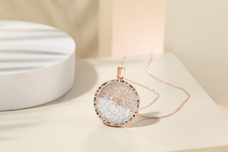 May include: Rose gold pendant necklace with a circular design. The pendant features a ring of colorful gemstones surrounding a detailed central design with clear crystals. The necklace is displayed on a neutral background.