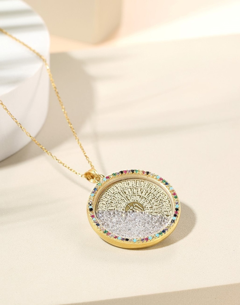 May include: A gold-colored pendant necklace with a round, ornate pendant. The pendant features a detailed design with colorful gemstones around the edge and clear crystals inside. The necklace chain is also gold-colored.