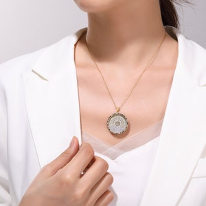 May include: A gold-toned necklace with a circular pendant featuring a detailed, ornate design. The pendant hangs from a delicate chain. The person is wearing a white blazer.