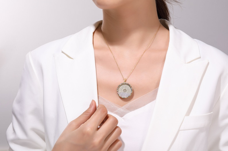 May include: A gold-toned necklace with a circular pendant featuring a detailed, ornate design. The pendant hangs from a delicate chain. The person is wearing a white blazer.