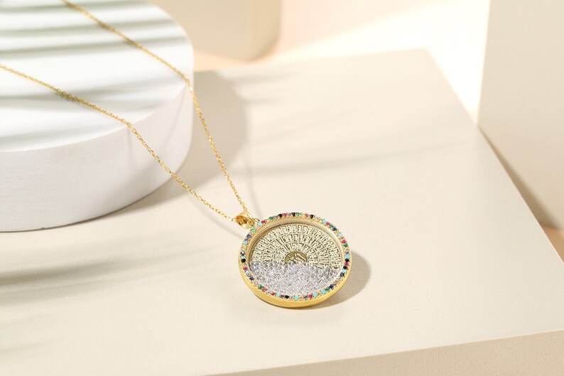 May include: A gold-colored pendant necklace with a round, detailed pendant. The pendant has a gold frame with a ring of colorful gemstones. The pendant's center features a detailed design with a silver and gold color scheme. The necklace chain is also gold.