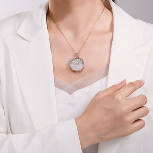 May include: A rose gold necklace with a round pendant. The pendant features a detailed design with clear and blue gemstones. The necklace is worn over a white top and a white blazer.
