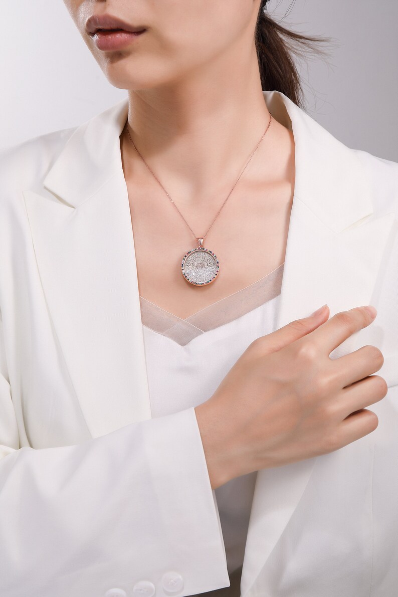 May include: A rose gold necklace with a round pendant. The pendant features a detailed design with clear and blue gemstones. The necklace is worn over a white top and a white blazer.