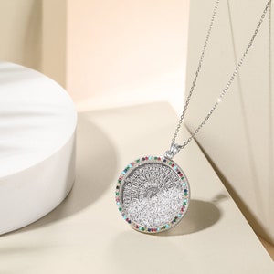 May include: A silver pendant necklace with a round, detailed pendant. The pendant has a central design surrounded by a ring of colorful gemstones. The necklace is displayed on a white surface.