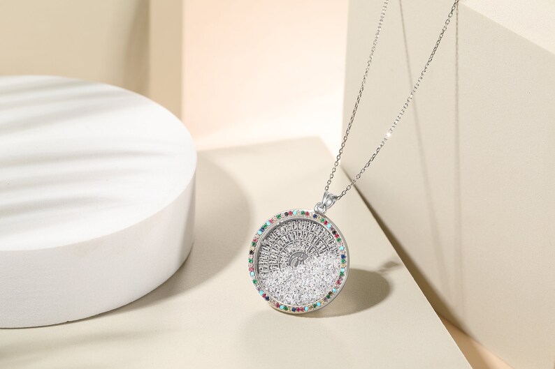 May include: A silver pendant necklace with a round, detailed pendant. The pendant has a central design surrounded by a ring of colorful gemstones. The necklace is displayed on a white surface.