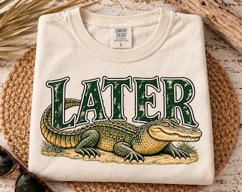May include: A cream-colored short-sleeve t-shirt featuring a graphic of a green alligator positioned below the word LATER in large, distressed dark green letters. The shirt has a Comfort Colors brand label at the inner neckline showing size L.
