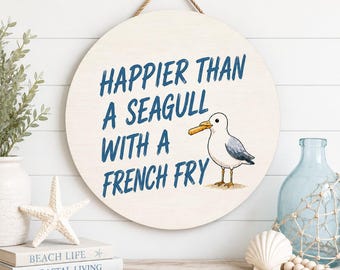 May include: Round wooden sign with the text "HAPPIER THAN A SEAGULL WITH A FRENCH FRY" in blue. A cartoon seagull with a french fry in its beak is to the right of the text. The sign is hanging on a white wall.