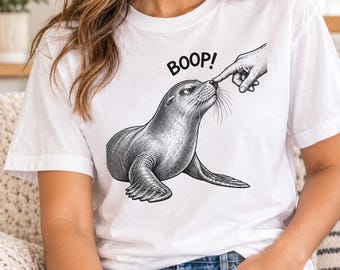 May include: A white short-sleeve t-shirt featuring a black and white ink-style illustration of a seal being touched on the nose by a human finger. Above the seal, the word BOOP! is printed in bold black capital letters in a simple font.