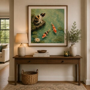 May include: A square framed wall art print featuring a calico cat sitting on a stone ledge overlooking a pond with two koi fish and a white water lily. The artwork is displayed above a wooden console table with a lamp, books, and a vase.