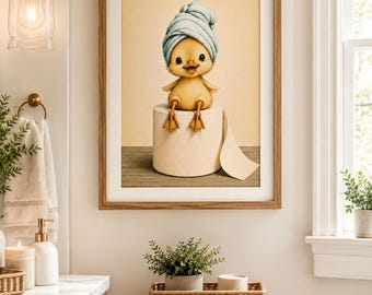 May include: A framed print featuring a yellow chick wearing a blue towel on its head, sitting on a roll of toilet paper. The artwork has a light brown wooden frame and is displayed in a bathroom setting.