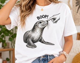 May include: A white t-shirt featuring a black and white line art illustration of a seal being touched on the nose by a human finger. Above the seal, the word BOOP! is printed in bold black capital letters. The design is centered on the chest.