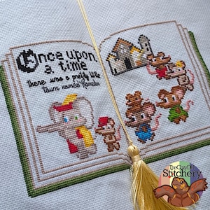 May include: A cross-stitch embroidery on light blue fabric depicting an open book with the text Once upon a time there was a pretty little town named Hamelin. The design features a gray elephant, a small mouse, and a castle with several mice.