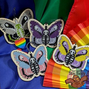 May include: Four butterfly-shaped wooden ornaments featuring cross-stitch patterns with skeleton-like designs in black, white, purple, blue, pink, and yellow. Each ornament has a dark string loop for hanging and a small rainbow heart charm.