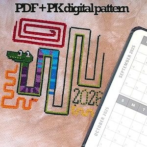 May include: Cross-stitch pattern featuring a colorful snake design with the text "2025". The design includes a green snake head, purple and blue body, and orange accents. A digital calendar is visible in the background.