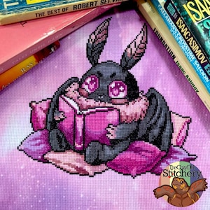 May include: A cross-stitch design featuring a cute, cartoon-style moth reading a book while resting on pillows. The moth is black with pink accents, and the book is bright pink. The background is a soft purple with books in the background.