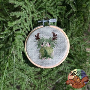 May include: A circular wooden embroidery hoop containing a cross-stitch design on grey fabric. The design features a small, green, furry creature with brown antlers, pink cheeks, and a light green body, centered within the hoop frame.