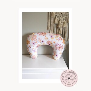 May include: A white nursing pillow with a floral pattern in shades of pink, orange, and yellow. The crescent-shaped pillow is displayed on a white surface. The Poppy Patterns PDF logo is in the bottom right corner.
