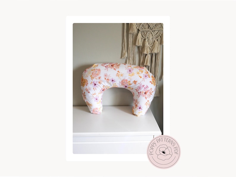 May include: A white nursing pillow with a floral pattern in shades of pink, orange, and yellow. The crescent-shaped pillow is displayed on a white surface. The Poppy Patterns PDF logo is in the bottom right corner.