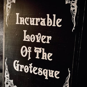 May include: A black wooden sign with white text that reads "Incurable Lover Of The Grotesque." The sign has decorative white corner accents. The sign is likely for home decor.