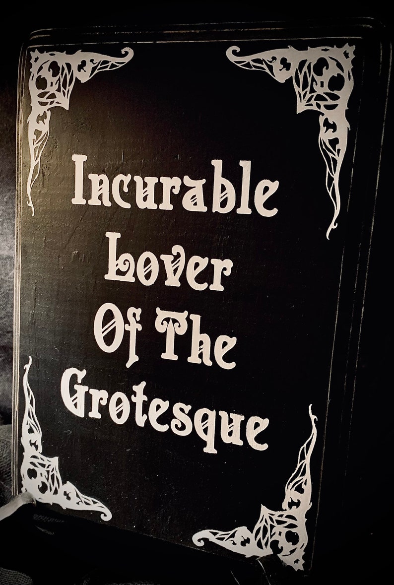 May include: A black wooden sign with white text that reads "Incurable Lover Of The Grotesque." The sign has decorative white corner accents. The sign is likely for home decor.