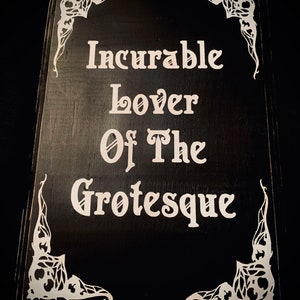 May include: A black wooden sign with white lettering that reads "Incurable Lover Of The Grotesque." The corners are decorated with white, ornate designs. The sign has a rustic, gothic aesthetic.
