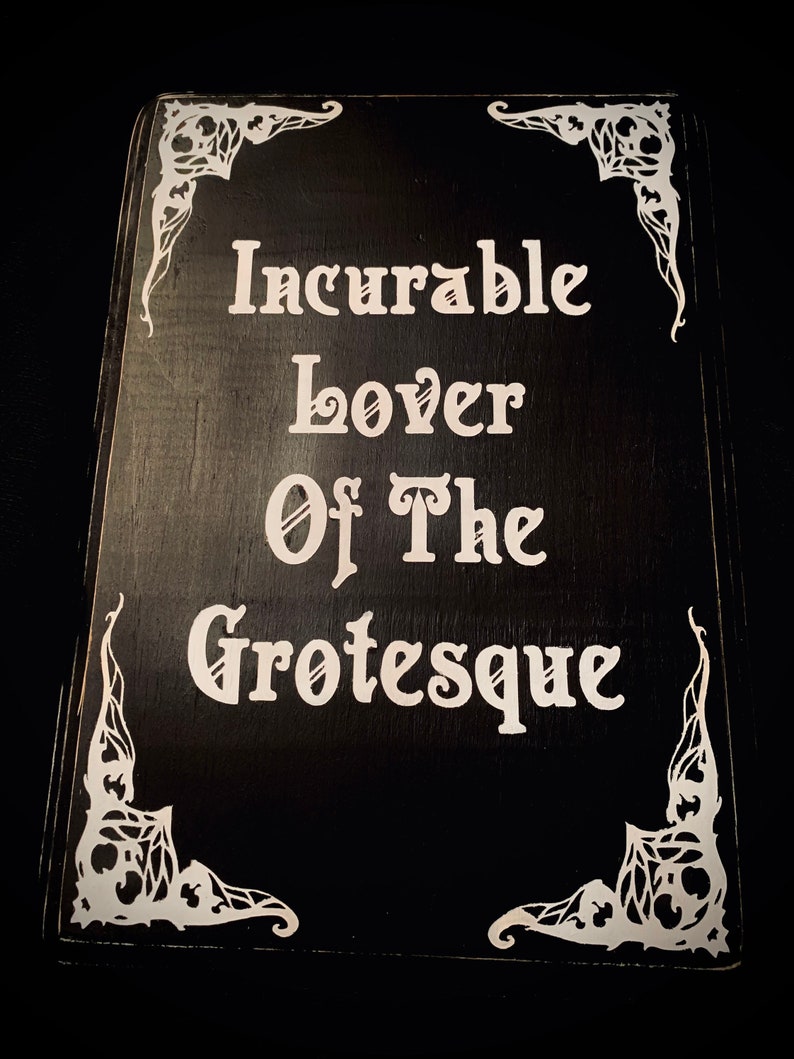 May include: A black wooden sign with white lettering that reads "Incurable Lover Of The Grotesque." The corners are decorated with white, ornate designs. The sign has a rustic, gothic aesthetic.