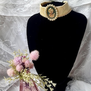 Può includere: Una collana girocollo in tessuto color crema con un ciondolo a cammeo ovale centrale raffigurante una figura di profilo color oro su sfondo verde scuro, circondato da un bordo di piccole perle bianche artificiali e rifinito con un bordo smerlato.