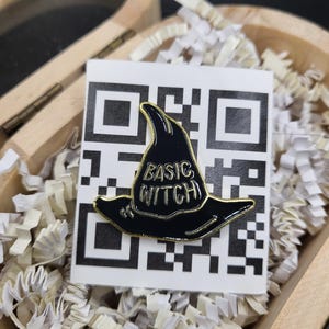Op de afbeelding: Zwarte emaille pin in de vorm van een heksenhoed met gouden rand. De woorden "BASIC WITCH" staan in witte letters op de hoed. De pin zit op een witte kaart met een QR-code, in een houten doos met papieren vulling.