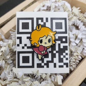 May include: A colourful enamel pin depicting a cartoon character with yellow hair and a red dress. The pin is set against a white QR code on a white background, inside a wooden box with white paper filler.