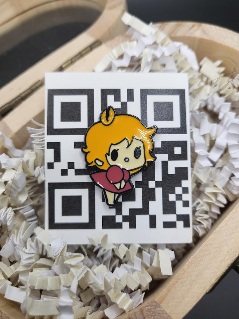 May include: A colourful enamel pin depicting a cartoon character with yellow hair and a red dress. The pin is set against a white QR code on a white background, inside a wooden box with white paper filler.