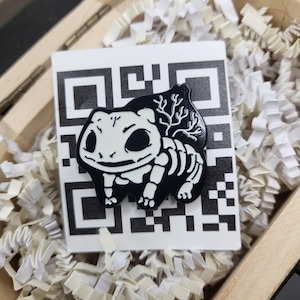 May include: A black and white enamel pin featuring a skeletal Bulbasaur design. The pin is set against a white QR code on a white background, nestled in a wooden box with paper filler.