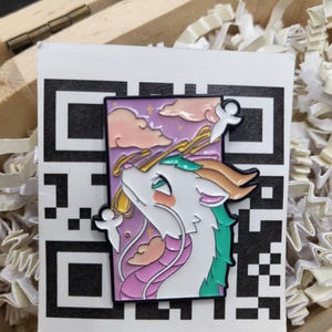 May include: A colorful enamel pin featuring a white dragon with green and orange accents, set against a purple sky with pink clouds and stars. The pin is on a white card with a black QR code.