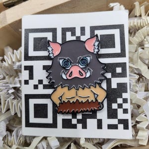 Puede incluir: Un colorido pin de esmalte con la imagen de una cabeza de jabalí de dibujos animados con una expresión feroz. El pin tiene una cabeza gris, orejas y hocico rosas y un torso marrón. Se coloca sobre una tarjeta blanca con un diseño de código QR negro.