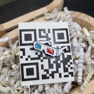 May include: A white enamel pin of 3D glasses with red and blue lenses, resting on a white card with a black QR code. The pin is in a wooden box with white paper filler.