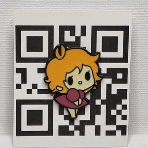 May include: A colourful enamel pin depicting a cartoon girl with orange hair and a pink dress. The pin is set against a white background with a black and white QR code design. A cute accessory.