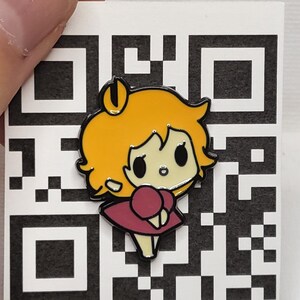 May include: A colourful enamel pin depicting a cartoon girl with yellow hair and a maroon dress. The pin has a black outline and is set against a white background with a QR code pattern. The pin is held by a person's fingers.