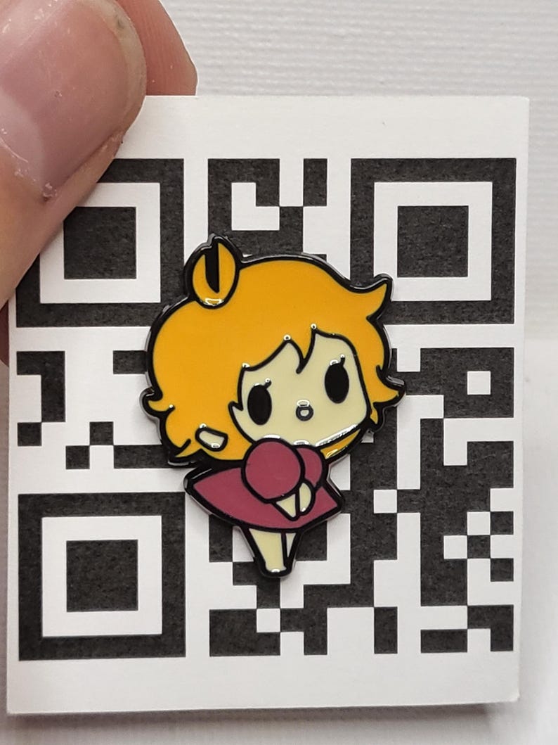 May include: A colourful enamel pin depicting a cartoon girl with yellow hair and a maroon dress. The pin has a black outline and is set against a white background with a QR code pattern. The pin is held by a person's fingers.