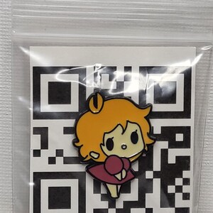 May include: A cartoon character enamel pin with orange hair and a maroon dress. The pin is set against a black and white QR code background, enclosed in a clear plastic bag.