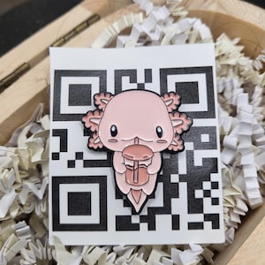 Könnte beinhalten: Ein rosa Emaille-Pin mit einem Axolotl, das ein Getränk hält, vor einem QR-Code auf weißem Hintergrund. Der Pin hat schwarze Umrisse und befindet sich in einer Holzkiste mit weißen Papierschnipseln.