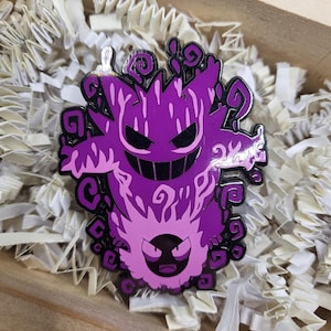 May include: A purple and black enamel pin featuring a stylized ghost design. The pin has a detailed design with a smiling face and swirling accents. The pin is set against a background of white paper shreds inside a wooden box.