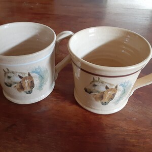 May include: Two ceramic mugs with horse head designs. The mugs are beige with brown and blue accents. One mug has a white horse and a brown horse, while the other mug has a white horse and a brown horse.