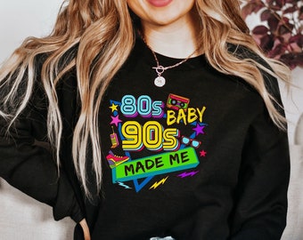 May include: Black sweatshirt with a colorful graphic design that says "80s Baby 90s Made Me". The graphic includes retro icons like a cassette tape, sunglasses, and a boombox.