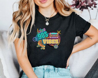 May include: A black t-shirt with a graphic design featuring the text "90s Vibes" in a colorful retro style. The design includes a roller skate, a cassette tape, and a Rubik's Cube.