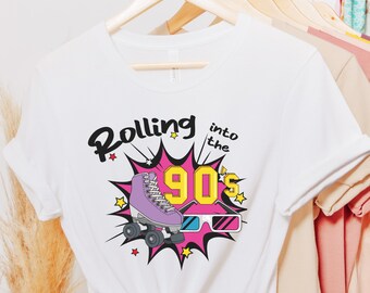 May include: White t-shirt with a colorful graphic design. The design features a purple roller skate, 3D glasses, and the text "Rolling into the 90's" in black and yellow. The shirt is hanging on a wooden hanger.