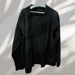 May include: A black knitted cardigan with a long sleeve and a v-neck. The cardigan is open in the front and has a ribbed texture.
