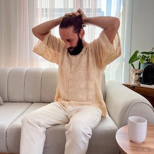 K&ouml;nnte beinhalten: Ein Mann in einem hellbeigen H&auml;kelhemd mit wei&szlig;em Knopfkragenshirt und wei&szlig;er Hose sitzt auf einem hellgrauen Sofa. Er zieht sich die Haare zu einem Knoten zusammen.