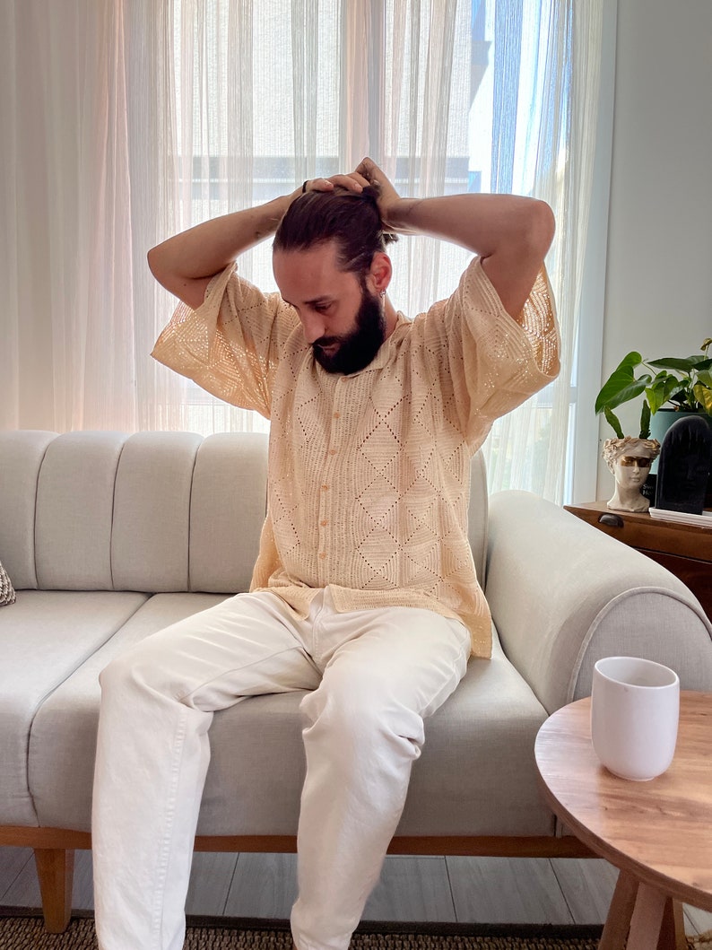 K&ouml;nnte beinhalten: Ein Mann in einem hellbeigen H&auml;kelhemd mit wei&szlig;em Knopfkragenshirt und wei&szlig;er Hose sitzt auf einem hellgrauen Sofa. Er zieht sich die Haare zu einem Knoten zusammen.